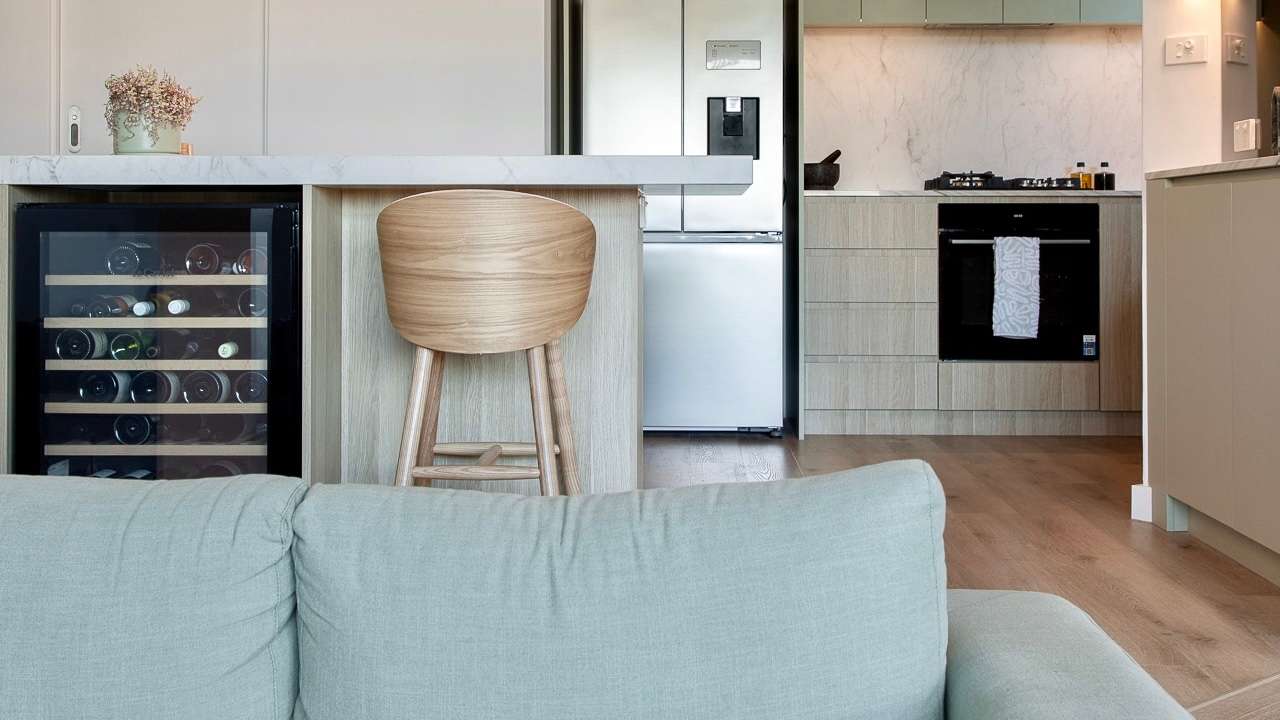 A living space with a sage-green sofa looking through to a kitchen with timber joinery and a wine fridge.